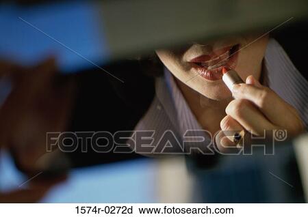 Stock Image - Young woman applying lipstick in a car rear view mirror. Fotosearch