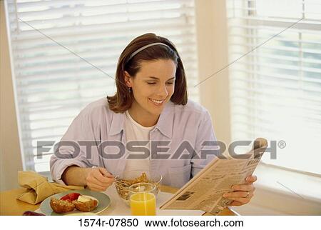 Stock Image - Young woman eating breakfast while reading a newspaper. Fotosearch