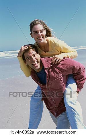 Stock Image - Young woman riding piggyback on a young man at the beach. Fotosearch