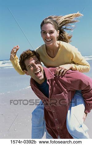 Stock Image - Young woman riding piggyback on a young man at the beach. Fotosearch