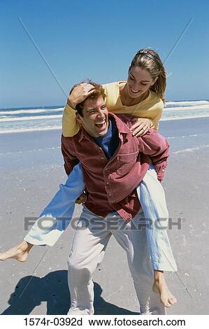 Stock Image - Young woman riding piggyback on a young man at the beach. Fotosearch