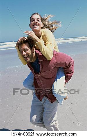 Stock Image - Young woman riding piggyback on a young man at the beach. Fotosearch