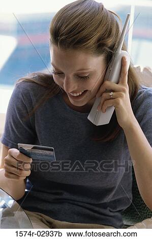 Stock Image - Young woman using a cordless phone holding a credit card. Fotosearch
