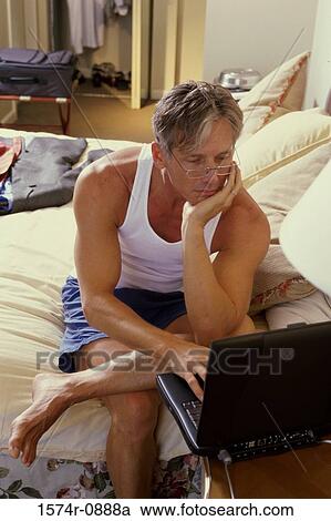 Stock Image - Businessman sitting on a bed in a hotel room working on a laptop. Fotosearch