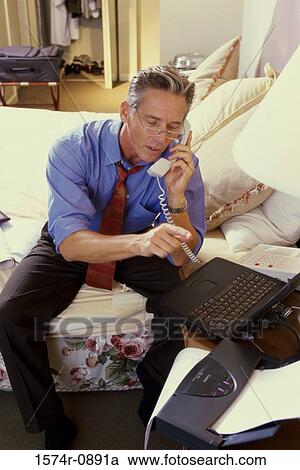Stock Image - Businessman sitting on a bed in a hotel room talking on a telephone. Fotosearch