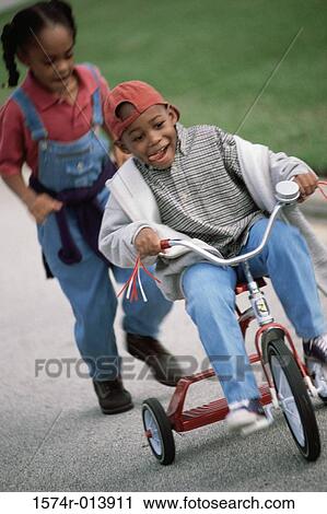 Stock Image - Close-up of a boy riding a tricycle with a girl running behind him. Fotosearch
