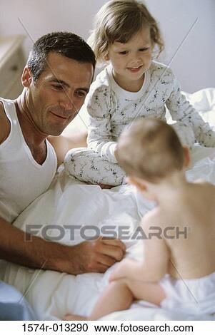 Stock Image - Father sitting with his son and daughter on a bed. Fotosearch