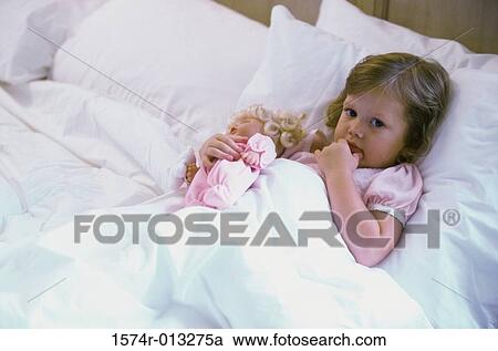 Stock Image - High angle view of a baby girl with a doll lying in bed. Fotosearch