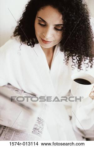 Picture - High angle view of a young woman holding a cup of coffee while reading a newspaper. Fotosearch