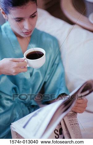 Stock Image - High angle view of a young woman holding a cup of coffee while reading a newspaper. Fotosearch