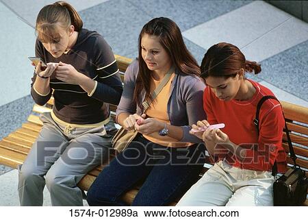Stock Image - High angle view of three teenage girls sitting on a bench applying make-up. Fotosearch