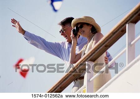 Stock Image - Low angle view of a mature woman taking a photograph with a mature man waving beside her on a cruise ship. Fotosearch