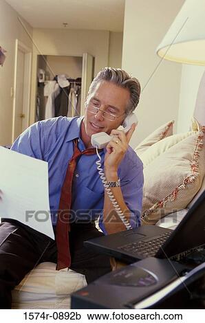 Stock Image - Mature man sitting on the bed and talking on a landline phone. Fotosearch