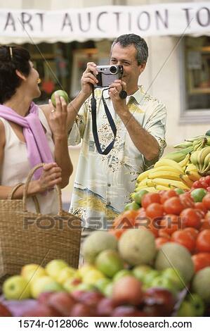 Stock Image - Mature man taking a photograph of a mature woman in front of a market stall. Fotosearch