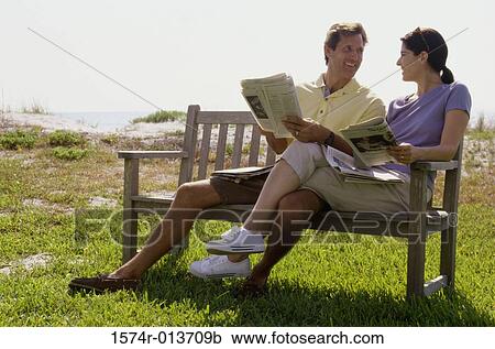 Stock Image - Mid adult couple sitting on a bench holding newspapers. Fotosearch
