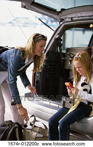Stock Image - Mother loading luggage in a car while her daughter plays a video game. Fotosearch