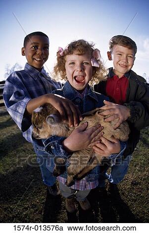 Stock Image - Portrait of a girl holding a kid goat in her arms with two boys standing beside her. Fotosearch