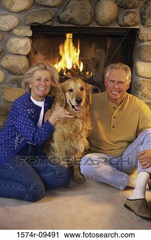 Stock Image - Portrait of a senior couple in front of a fireplace with a dog. Fotosearch