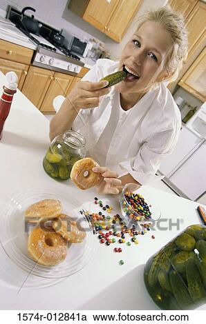 Stock Image - Portrait of a young woman eating a pickle and holding a donut. Fotosearch