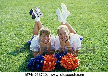 Stock Image - Portrait of two cheerleaders lying down on a field with pom-poms in front of them. Fotosearch