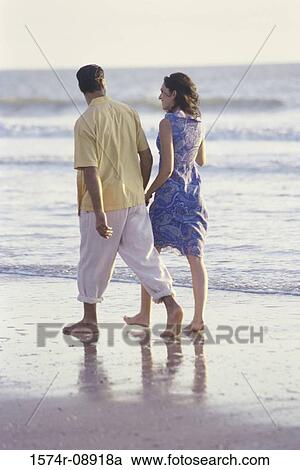 Stock Image - Rear view of a young couple walking on the beach. Fotosearch