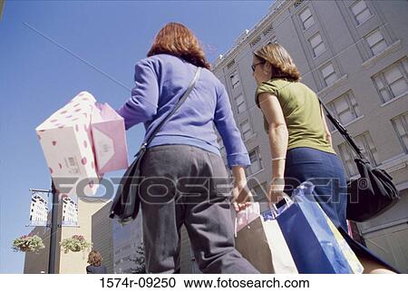 Stock Image - Rear view of two young women walking carrying shopping bags. Fotosearch