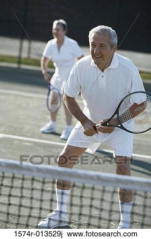 Stock Image - Senior man playing tennis with a senior woman in the background. Fotosearch