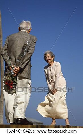 Stock Image - Senior man standing in front of a senior woman with a bouquet of flowers behind his back. Fotosearch