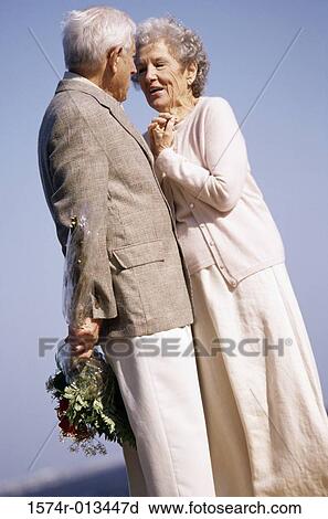 Stock Image - Senior man standing in front of a senior woman with a bouquet of flowers behind his back. Fotosearch