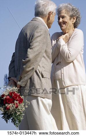 Stock Image - Senior man standing in front of a senior woman with a bouquet of flowers behind his back. Fotosearch