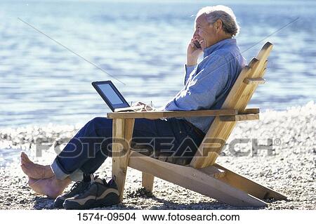 Picture - Side profile of a senior man sitting on the beach talking on a mobile phone. Fotosearch