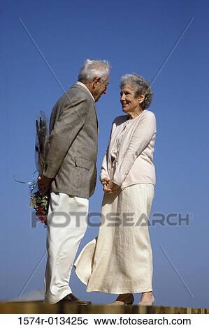 Stock Image - Side profile of a senior man standing in front of a senior woman with a bouquet of flowers behind his back. Fotosearch