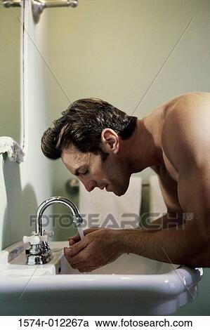 Stock Image - Side profile of a young man washing his face in a bathroom sink. Fotosearch