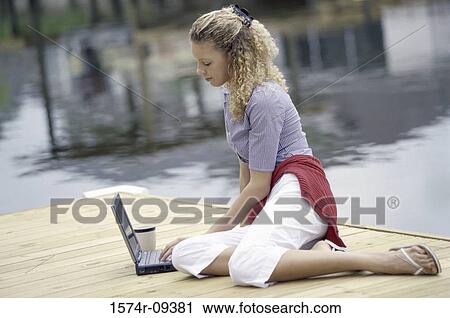 Stock Image - Side profile of a young woman sitting on a pier using a laptop. Fotosearch