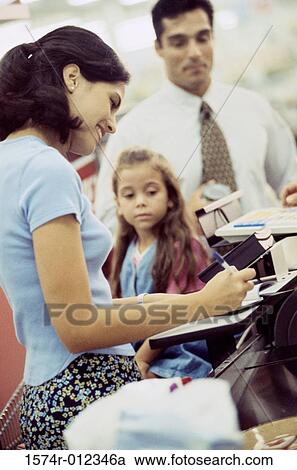 Stock Image - Side profile of a young woman with her daughter standing at a checkout counter in a supermarket. Fotosearch