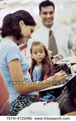 Stock Image - Side profile of a young woman with her daughter standing at a checkout counter in a supermarket. Fotosearch