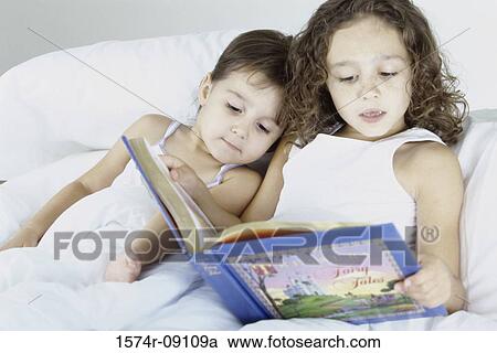 Stock Image - Two sisters lying in bed together reading a book. Fotosearch