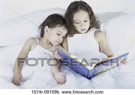 Stock Image - Two sisters lying in bed together reading a book. Fotosearch