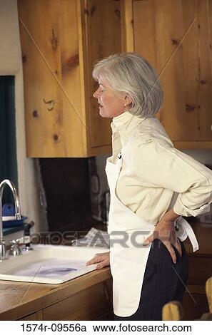 Woman standing in a kitchen holding her back in pain View Large Photo Image Stock Image - Woman standing in a kitchen holding her back in pain. Fotosearch