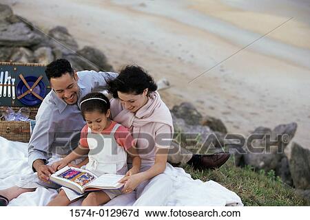 Stock Image - Young couple and their daughter reading a book on the beach. Fotosearch