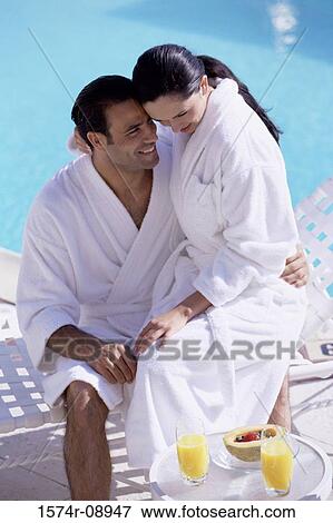 Stock Photo - Young couple sitting together at a swimming pool wearing bathrobes. Fotosearch