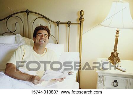 Stock Photo - Young man holding a newspaper and laughing in bed. Fotosearch