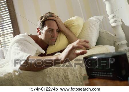 Stock Image - Young man lying on a bed reaching for the alarm clock. Fotosearch