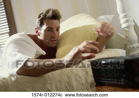 Stock Image - Young man lying on a bed reaching for the alarm clock. Fotosearch