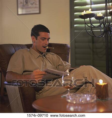 Stock Image - Young man sitting on a couch reading a book and smoking a cigar. Fotosearch