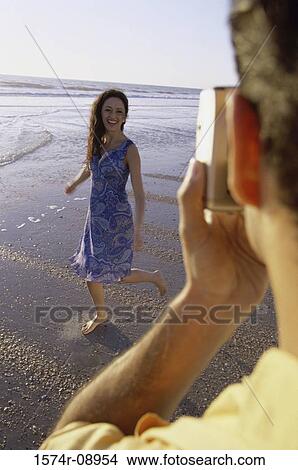 Picture - Young man taking a photograph of a young woman at the beach. Fotosearch