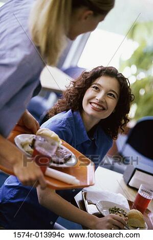 Young woman holding a tray of food looking at a woman seated at a table View Large Photo Image Stock Image - Young woman holding a tray of food looking at a woman seated at a table. Fotosearch