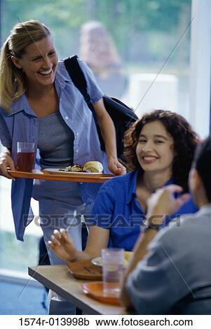 Young woman holding a tray of food standing behind a woman seated at a table View Large Photo Image Stock Image - Young woman holding a tray of food standing behind a woman seated at a table. Fotosearch