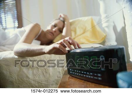 Stock Photography - Young woman lying in bed reaching for an alarm clock. Fotosearch
