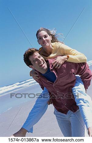 Stock Image - Young woman riding piggyback on a young man on the beach. Fotosearch